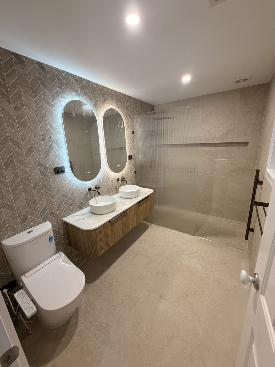 Same ensuite after renovation, with herringbone tiles, backlit mirrors and walk-in shower